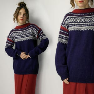 Puede incluir: Un suéter azul marino con cuello y puños rojos. El suéter presenta un patrón geométrico en blanco y gris en el pecho y la parte superior de las mangas. Las mangas son largas y el suéter parece estar hecho de un material suave y cálido.