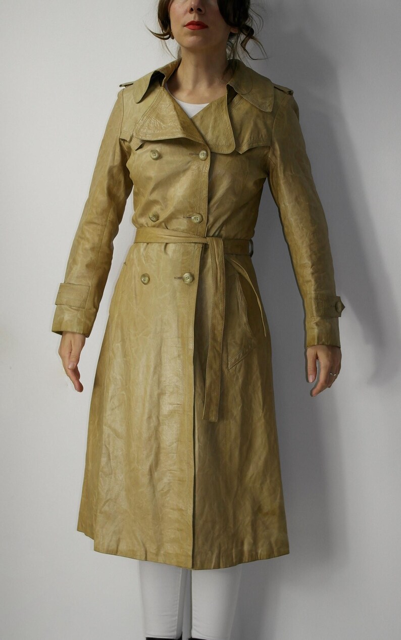 May include: A long, tan leather trench coat with a double-breasted closure, a belt, and a collar.