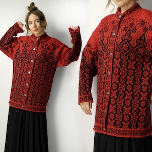 May include: A red cardigan with a black floral pattern. The button-down sweater has long sleeves and a high collar. The intricate design covers the entire garment, creating a striking visual effect. The cardigan is worn over a black pleated skirt.