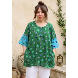 Puede incluir: Una túnica verde con un estampado de círculos azules, morados y amarillos. La túnica tiene mangas azules y una botonadura frontal. La modelo lleva pantalones blancos.