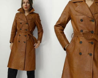 May include: A brown leather trench coat with a double-breasted design, a belt, and large lapels. The coat has long sleeves and a knee-length hem. The coat is worn by a person wearing black pants.