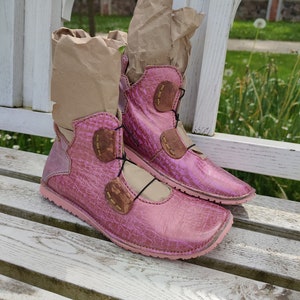 May include: A pair of pink leather ankle boots with brown leather accents and laces. The boots are sitting on a white wooden bench.