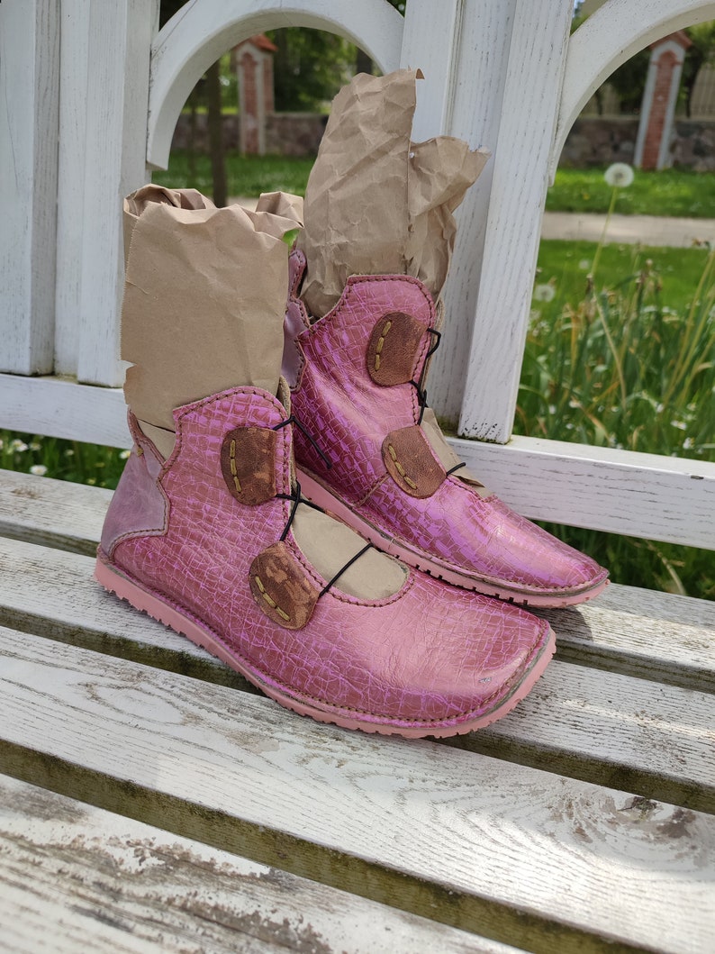 May include: A pair of pink leather ankle boots with brown leather accents and laces. The boots are sitting on a white wooden bench.