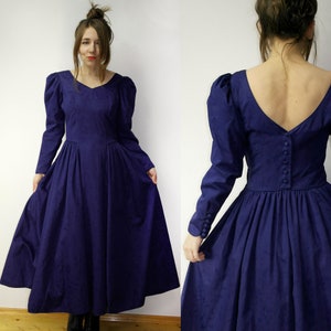 May include: A long-sleeved, dark purple dress with a gathered skirt and a low, V-shaped back. The dress has puffed sleeves and a button closure down the back.