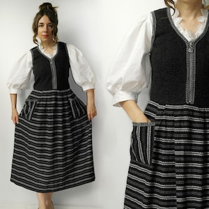 Op de afbeelding: Een zwart-wit gestreepte jurk met een donkergrijze vest en witte blouse. De jurk heeft een V-hals vest met een rits en zakken. De mouwen zijn pofmouwen en de rok heeft horizontale strepen.