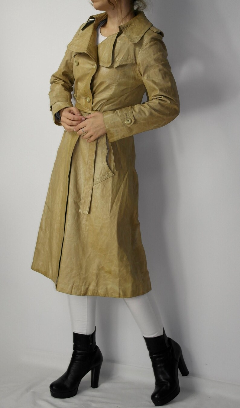May include: A long, tan leather trench coat with a double-breasted closure, a belt, and a collar that can be worn up or down.