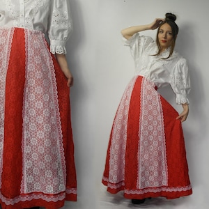 Può includere: Una gonna lunga rossa con pannelli e finiture in pizzo bianco, abbinata a una camicetta bianca traforata. La gonna ha una silhouette ampia e fluida, e i dettagli in pizzo aggiungono un delicato contrasto al tessuto rosso vibrante. La camicetta ha una chiusura a bottoni e maniche a balze.