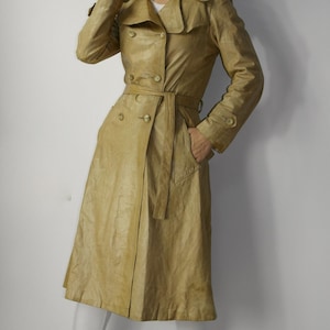 May include: A long, beige leather trench coat with a double-breasted closure, a belt, and pockets. The coat is worn by a person with brown hair and red lipstick.