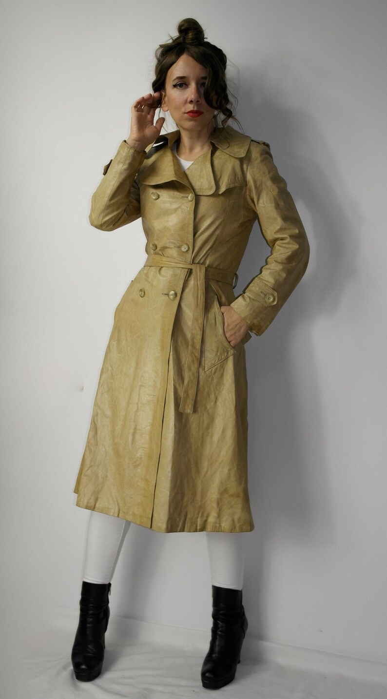 May include: A long, beige leather trench coat with a double-breasted closure, a belt, and pockets. The coat is worn by a person with brown hair and red lipstick.