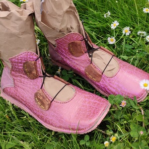 May include: Two pink leather boots with brown leather accents and laces. The boots are sitting on a bed of green grass and white flowers.