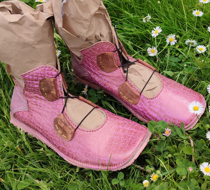 May include: Two pink leather boots with brown leather accents and laces. The boots are sitting on a bed of green grass and white flowers.
