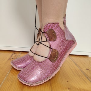 May include: Pink leather shoes with black laces and brown leather accents. The shoes have a unique design with a cut-out detail on the top.