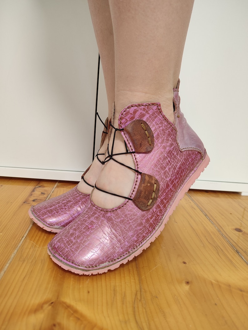 May include: Pink leather shoes with black laces and brown leather accents. The shoes have a unique design with a cut-out detail on the top.