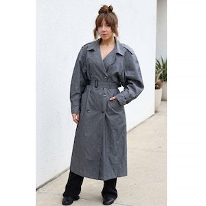 May include: A long, double-breasted trench coat in a muted charcoal grey colour. The coat features a wide notched collar, shoulder epaulets, long sleeves with buckled cuffs, and a matching waist belt with a rectangular buckle. It is worn over black trousers.