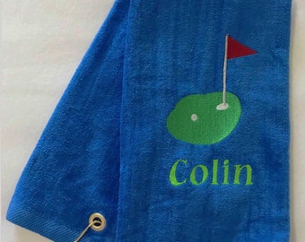 May include: Blue golf towel with a green embroidered golf course design, a red flag, and the name "Colin" in green. The towel has a loop and a metal ring for hanging.