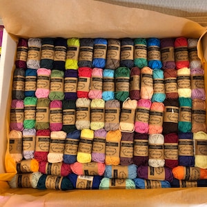 Puede incluir: Una caja llena de ovillos de lana de colores, cada uno etiquetado con "Catona". La lana está dispuesta en filas y columnas, con una variedad de colores, incluyendo rojo, azul, verde, amarillo y morado.