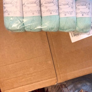 May include: A pack of five skeins of light teal yarn. Each skein has a label with the text "Organicon Scheepjes Premium Blend". The yarn is packaged in clear plastic and sits inside a brown cardboard box.