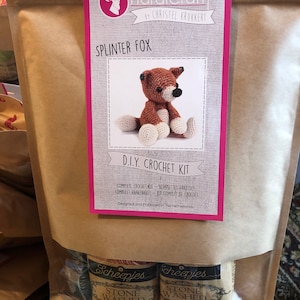May include: A brown paper bag with a clear window displaying a DIY crochet kit. The kit includes yarn and instructions to create a crocheted fox. The kit is labeled "Splinter Fox" and "D.I.Y. Crochet Kit". The yarn is labeled "Stone Washed".