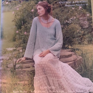 Puede incluir: Una portada de revista para Rowan Knitting & Crochet, número 43. La portada presenta a una mujer con un suéter de punto azul claro y una falda de encaje blanca. La mujer está sentada en un banco de piedra en un jardín.