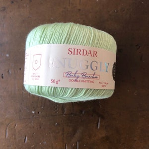 Puede incluir: Un ovillo de hilo de color verde claro etiquetado como "Sirdar Snuggly Baby Bamboo Double Knitting" con un peso de 50 gramos y una longitud de 95 metros o 104 yardas.