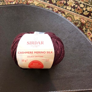 Pode incluir: Uma bola de fio Sirdar Cashmere Merino Silk de cor bordô escura. O rótulo diz "Sirdar West Yorkshire Est. 1900 Cashmere Merino Silk Double Knitting 50g* *116 m/127 yds approx West Yorkshire British Design Expertise and Craftsmanship"
