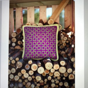 May include: A purple and green patterned throw pillow with a yellow crocheted border sits on a pile of stacked logs.