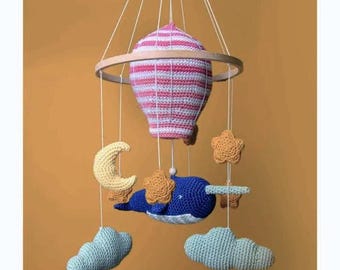 Puede incluir: Un móvil de bebé de crochet con un globo aerostático de rayas rosas y blancas, una narval azul, estrellas amarillas, una luna creciente amarilla y dos nubes verdes.