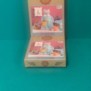 May include: Two open craft kit boxes with illustrations of a cat wearing a dress and knitting. The boxes are light brown with the text "Wool Mix Felt Craft Kit Mrs. Cat loves knitting". The background is teal.