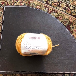Op de afbeelding: Een bol gele Sirdar Snuggly Cashmere Merino garen voor dubbel breien. Het etiket geeft 50g (1,75 oz) en 130m (142 yds) aan.