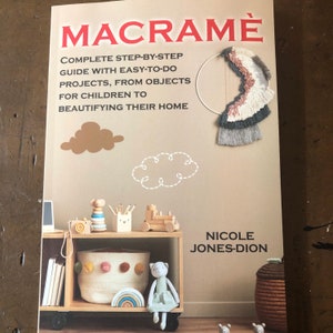 May include: A book titled "Macrame: Complete Step-by-Step Guide with Easy-to-Do Projects, From Objects for Children to Beautifying Their Home" by Nicole Jones-Dion. The cover features a macrame wall hanging and a child's room with a wooden shelf, toys, and a storage basket.