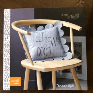 May include: A book titled "Felt Sew Good" with a gray felt pillow on a wooden chair. The book cover features a simple design with the title in black text and the words "30 Simple & Stylish Felt Projects". The book is part of the "Simple Makes" series.