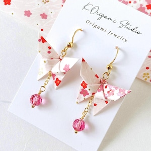 Könnte beinhalten: Origami-Schmetterlingsohrringe mit goldfarbenen Haken und Ketten. Die Papier-Schmetterlinge sind weiß mit einem rosa Blumenmuster und roten Akzenten. Jeder Ohrring hat eine rosa Kristallperle. Text: "K Origami Studio Origami Jewelry".
