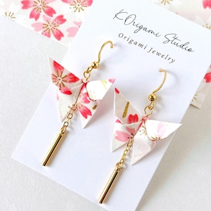 Könnte beinhalten: Origami-Schmetterlingsohrringe mit goldfarbenen Akzenten. Die Ohrringe zeigen Papier-Schmetterlinge mit einem rosa Blumenmuster und goldfarbenen Akzenten. Sie haben goldfarbene Ohrhaken und einen goldfarbenen Stäbchenanhänger.