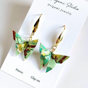 May include: A pair of origami butterfly earrings with gold-colored hooks. The butterfly wings are green, yellow, and brown. The earrings are displayed on a white card that reads "K Origami Studio Origami Jewelry". The card also says "Pierce" and "Clip-on".