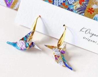 Può includere: Un paio di orecchini dorati a forma di uccello. Gli uccelli sono realizzati con carta origami colorata con motivi blu, viola e oro. Gli orecchini hanno un design delicato e artistico, perfetti per ogni outfit.