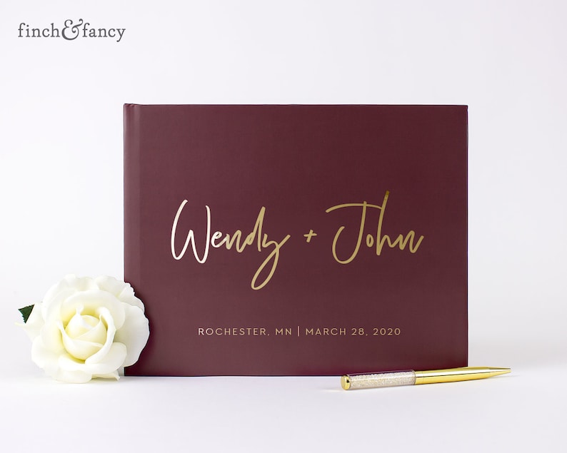 May include: Burgundy guest book with gold foil lettering. The names "Wendy + John" are written in cursive. The text "Rochester, MN | March 28, 2020" is printed below the names.