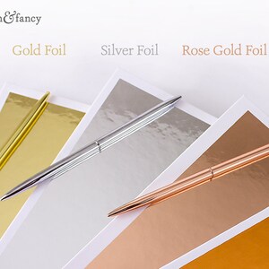 May include: Three pens with different colored foil finishes: gold, silver, and rose gold. The pens are lying on top of sheets of paper with the same colored foil finishes.