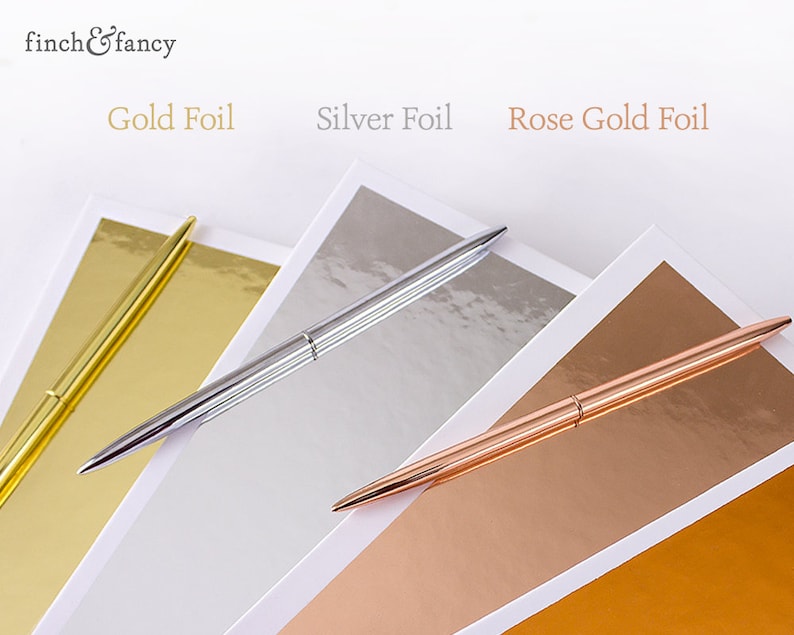 May include: Three pens with different colored foil finishes: gold, silver, and rose gold. The pens are lying on top of sheets of paper with the same colored foil finishes.