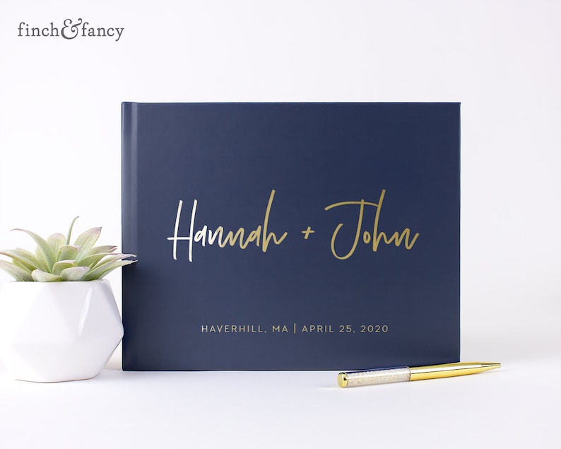 May include: A navy blue guest book with gold lettering that reads "Hannah + John" and "Haverhill, MA | April 25, 2020".