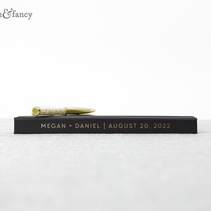 May include: A black guest book with gold lettering that reads "MEGAN + DANIEL | AUGUST 20, 2022". A gold pen with clear rhinestones is resting on top of the book.