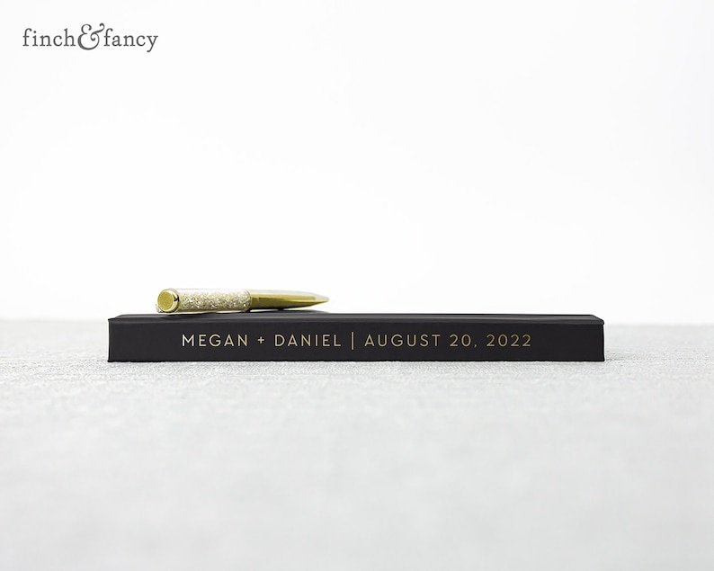 May include: A black guest book with gold lettering that reads "MEGAN + DANIEL | AUGUST 20, 2022". A gold pen with clear rhinestones is resting on top of the book.