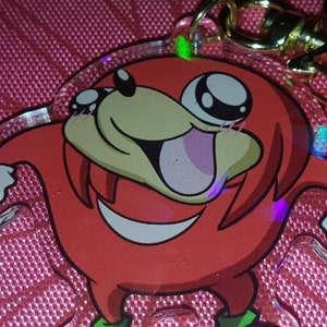 May include: A red and white cartoon character keychain with a gold star charm. The character has large eyes and a wide smile. The keychain is made of clear acrylic with a holographic finish.