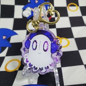 Peut inclure: Un porte-clés en acrylique transparent avec un fantôme de dessin animé portant un casque audio. Le fantôme a un corps blanc, un casque violet et un visage souriant. Le porte-clés a un fermoir doré et un charm en forme de cœur. Les bords sont violets pailletés.