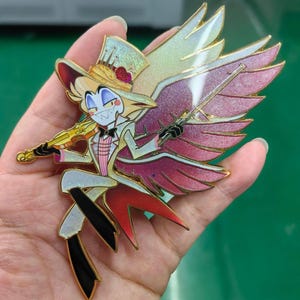 May include: A colorful enamel pin featuring a cartoon character with a top hat, bow tie, and wings, holding a violin. The pin has gold accents and a red, pink, and white color scheme. The character is smiling with sharp teeth.