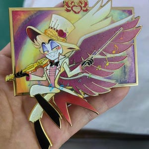 May include: A detailed enamel pin featuring a cartoon character with a top hat, wings, and a violin. The pin has a gold border and a colorful background. The character is dressed in a white suit with a pink vest and black accents.