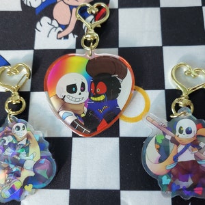 Op de afbeelding: Drie iriserende sleutelhangers met personages uit het populaire videospel "Undertale". De sleutelhangers zijn in de vorm van een hart, een halve maan en een cirkel. De personages zijn Ink Sans, Error Sans en een combinatie van de twee.