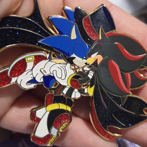 May include: A detailed enamel pin featuring Sonic the Hedgehog and Shadow the Hedgehog embracing. The pin has a gold outline, with Sonic in blue and white, and Shadow in black and red. The design includes glitter accents.