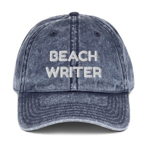 Op de afbeelding: Een blauwe denim baseballpet met de tekst "Beach Writer" geborduurd in wit op de voorkant.