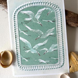 Puede incluir: Una impresi&oacute;n que representa una bandada de gaviotas blancas con picos rojos, sobre un fondo verde azulado dentro de un marco decorativo. La obra de arte est&aacute; en una tarjeta blanca, con conchas marinas y acentos de madera flotante.