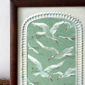 Puede incluir: Una obra de arte enmarcada que presenta una bandada de gaviotas blancas con picos rojos volando sobre un fondo verde claro. La obra est&aacute; encerrada en un borde blanco decorativo y un marco de madera marr&oacute;n oscuro, creando una est&eacute;tica vintage.